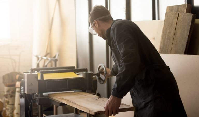 年轻的木匠或工匠穿着工作服和防护眼镜在小车间内木工，操作固定式动力刨床，加工修剪木板DeWalt DW735X刨床